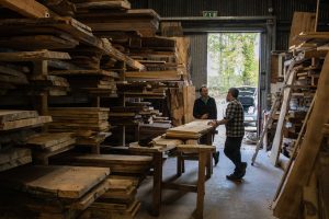 A customer stands inside the wood shed, talking to a Real Wood Studios maker, surrounded by shelves stacked with planks of beautiful wood.